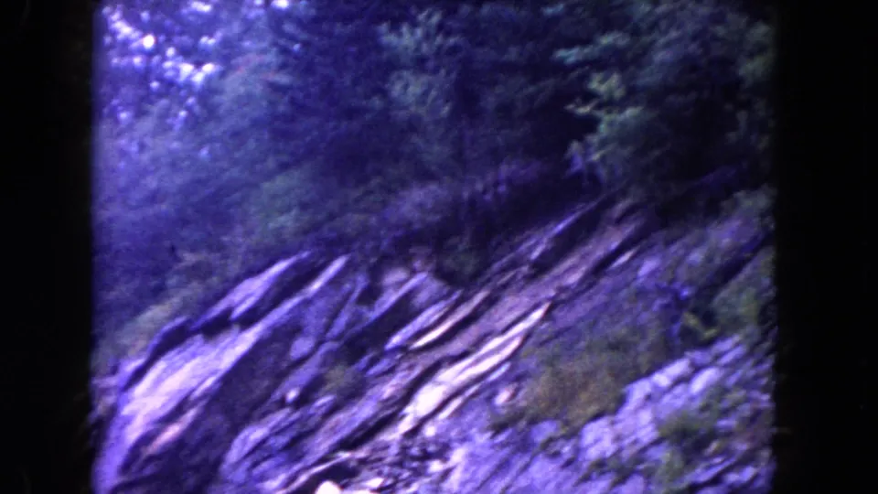 Aged 8mm mountain scenery, showing angled rock strata, evergreen growth, and a purple-blue film cast.