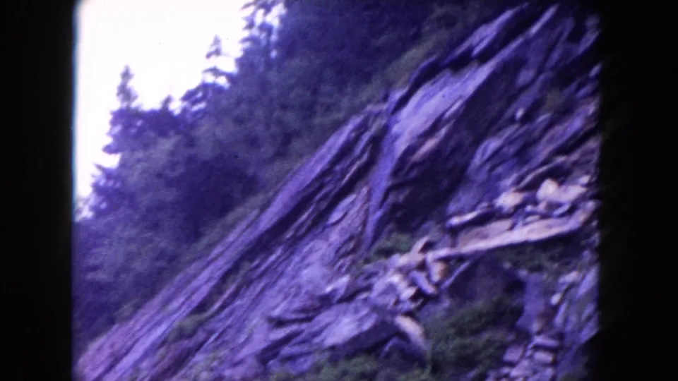 1960s 8mm roadside-style view of a steep rocky mountainside with trees and bright sky at the frame edge.