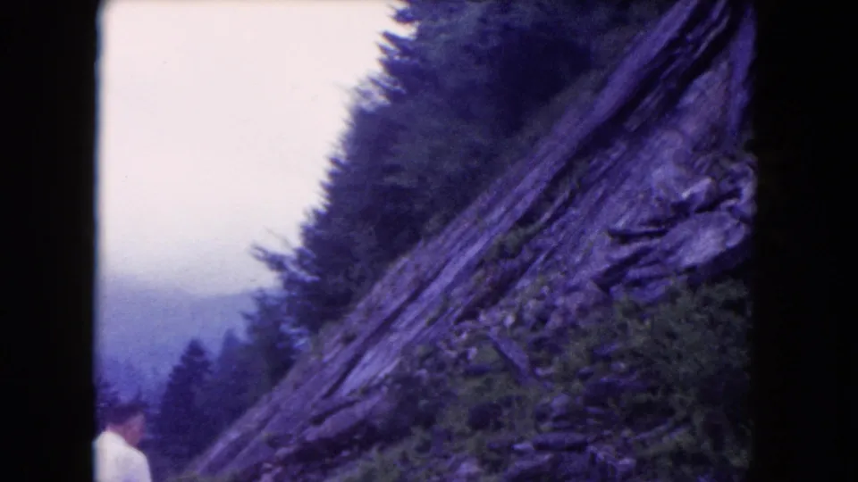 Aged 8mm frame of a rocky forested slope, hazy sky, and a partial person at the lower edge without an identifiable face.