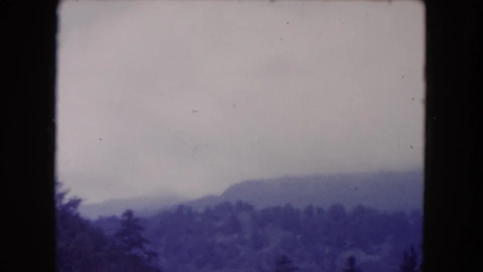 1960 8mm home-movie view of a hazy wooded valley and rounded ridge beneath an overcast sky.
