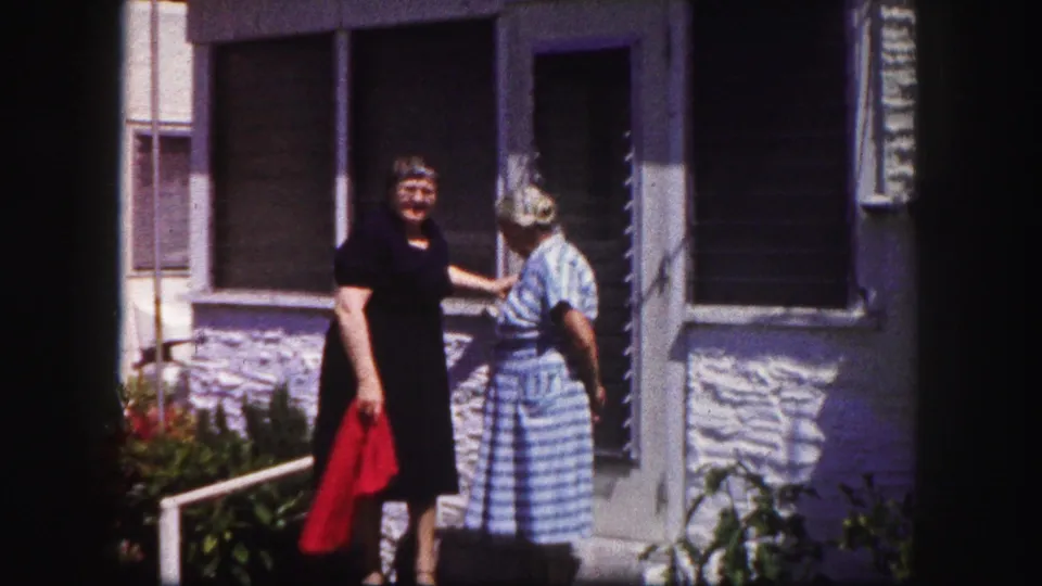 Two older women in 1960 Tampa home-movie footage stand by a stucco house, one holding a red cloth.