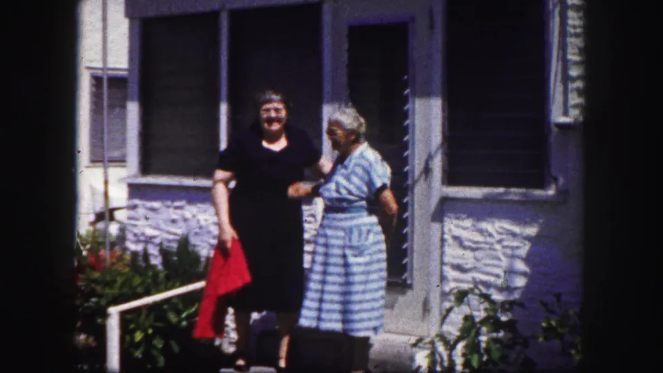 Two older women outside a Tampa residence in 1960, with stucco wall, louvered windows, plants, and a red cloth visible.
