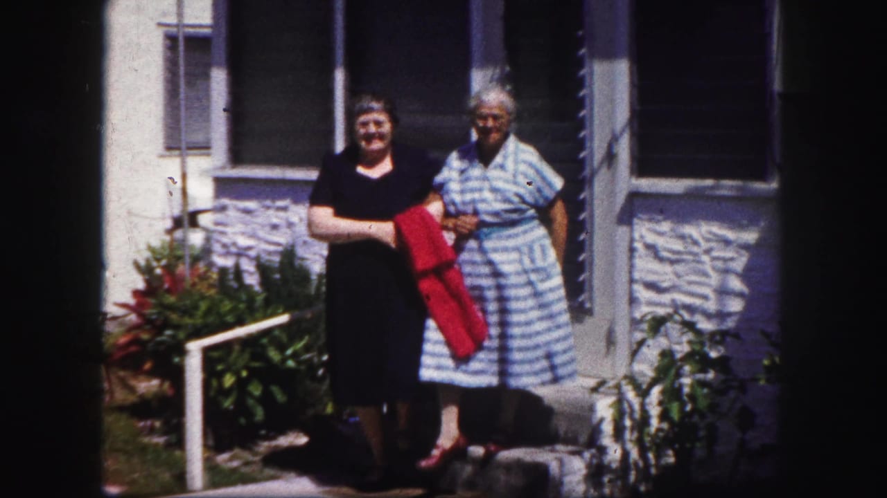 Two women in dresses pause or walk slowly outside a Tampa residence, with porch windows and greenery behind them.