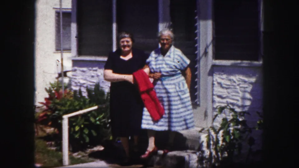 Two women in dresses pause or walk slowly outside a Tampa residence, with porch windows and greenery behind them.