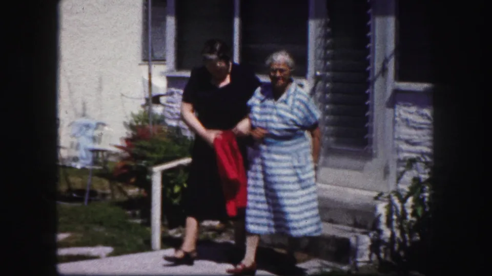 The pair continue arm in arm along the residential sidewalk, framed by porch steps and a sunlit house exterior.
