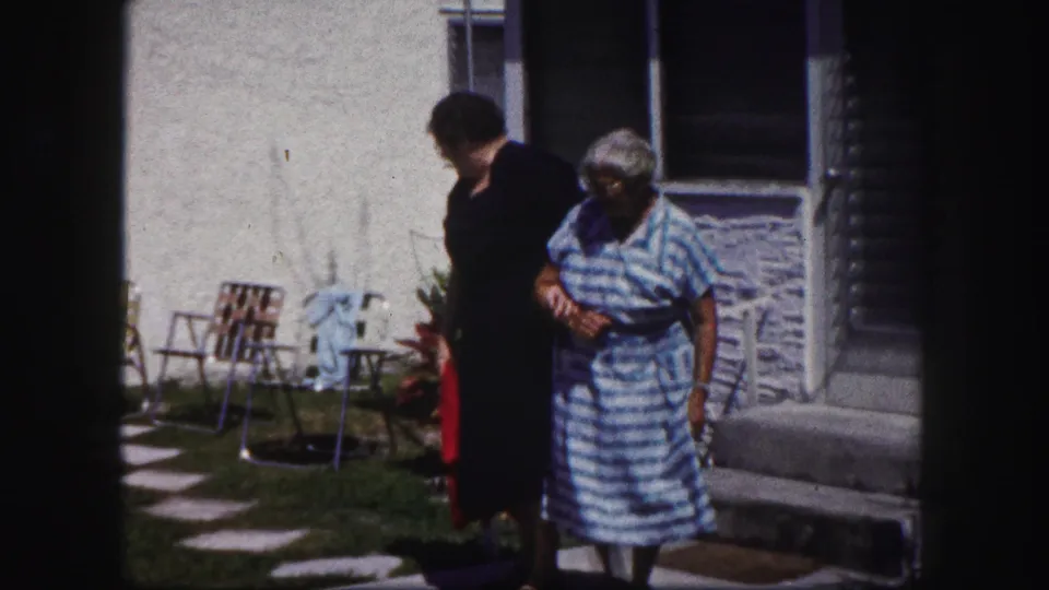 Two women move along the sidewalk outside the house, with yard chairs and porch details visible in the background.