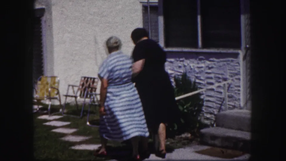 A younger woman in black assists an older woman in a striped dress along a yard path outside a Tampa house in 1960 footage.