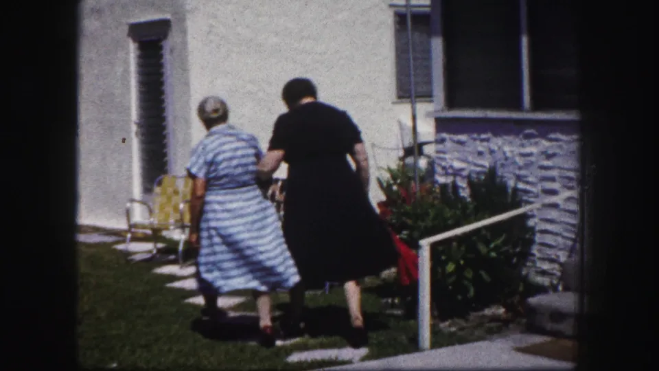 The pair move away from the camera along a stepping-stone path, with yard chairs and house siding visible behind them.