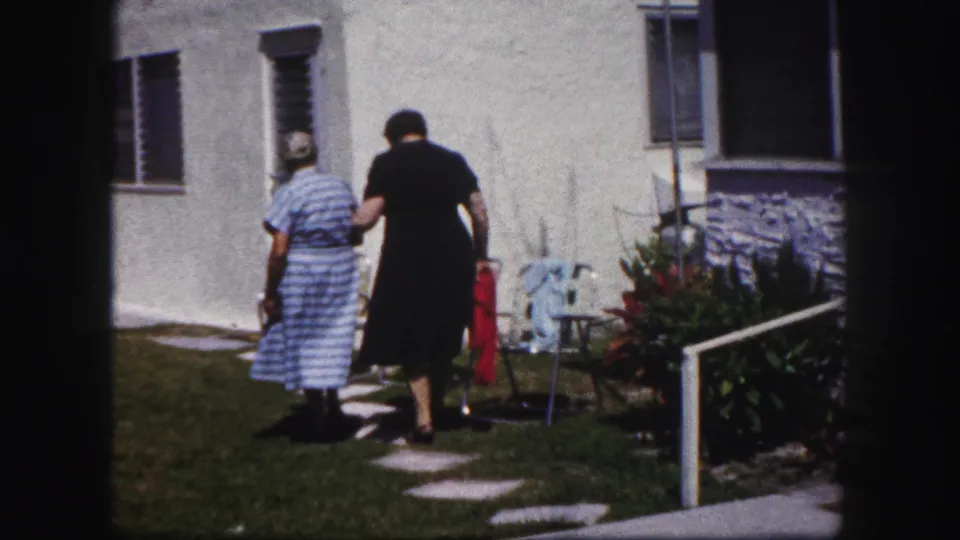 The younger woman walks beside the older woman on the path, moving away from camera in a 1960 Tampa backyard scene.