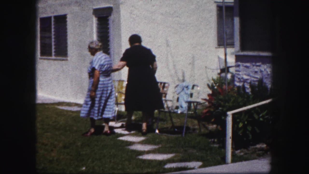 Two women stand on a lawn near patio chairs outside a white stucco house.