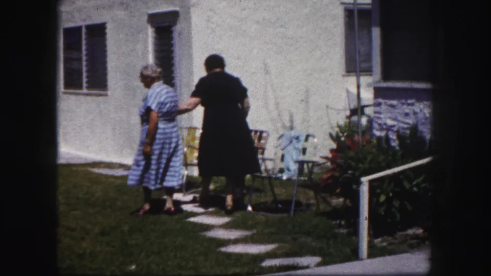In 1960 Tampa home-movie footage, two adult women stand near patio chairs and stepping stones outside a white stucco residence.