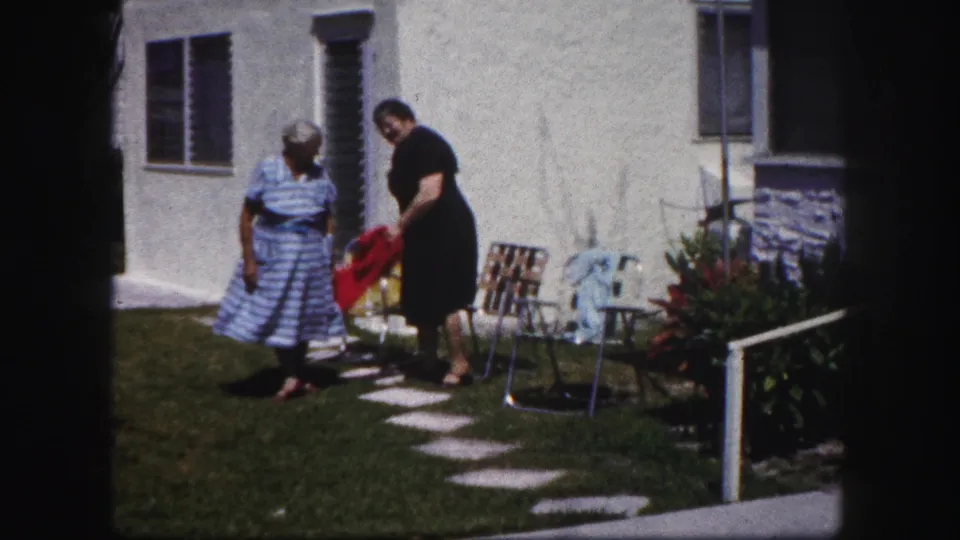 The dark-dressed woman handles a red cloth or bag while the striped-dress woman stands nearby in the Tampa yard.