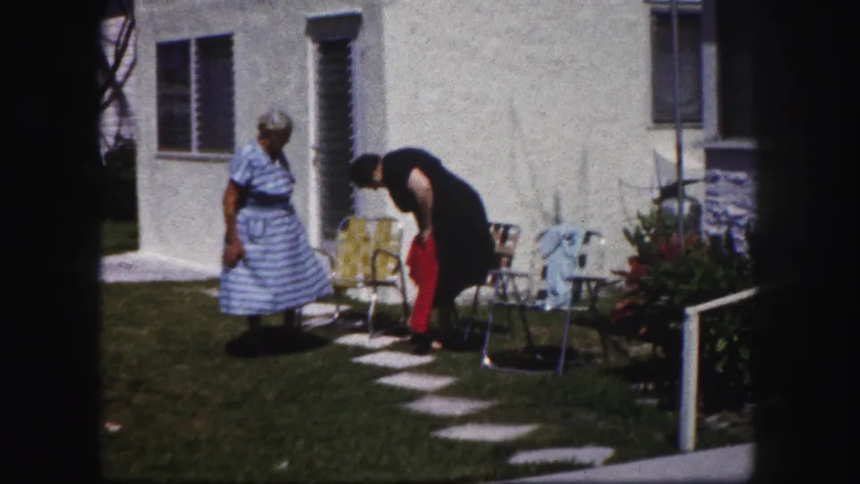 One adult woman bends to adjust a red item on the lawn as another watches near mid-century patio chairs outside the house.
