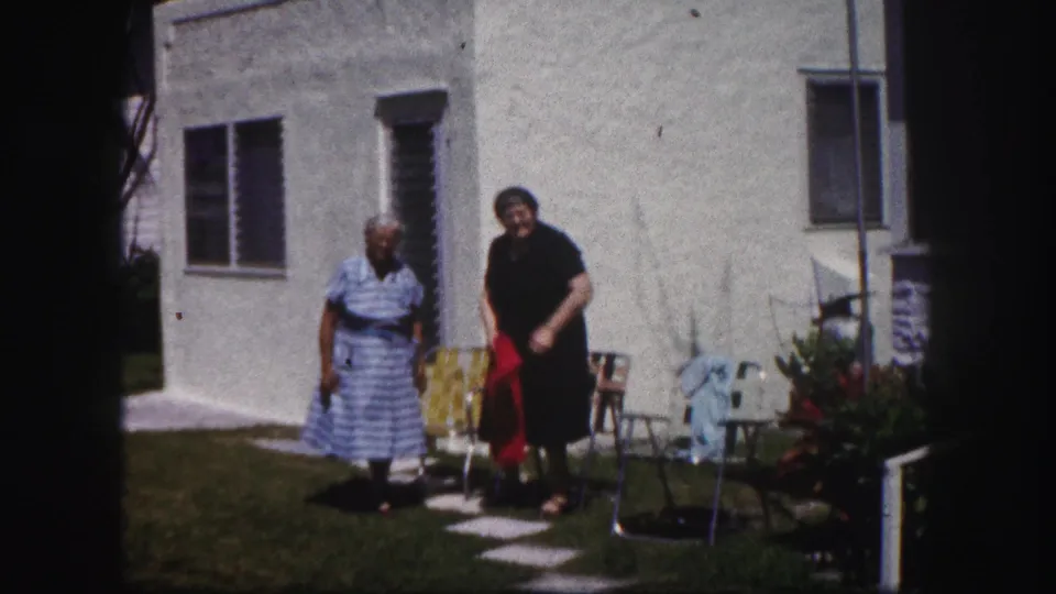 Two adult women face the camera in a Tampa residential yard, with a red item and patio chairs beside the white stucco house.