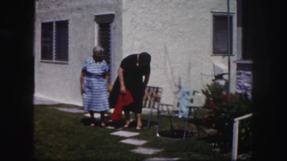 In 1960 Tampa home-movie footage, two older women in dresses stand by lawn chairs beside a white stucco house and lawn.