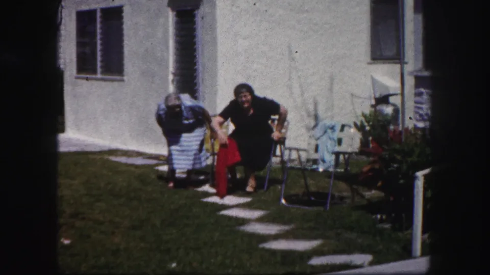 Two older women in dresses move toward metal lawn chairs along a stepping-stone path in a Tampa backyard.