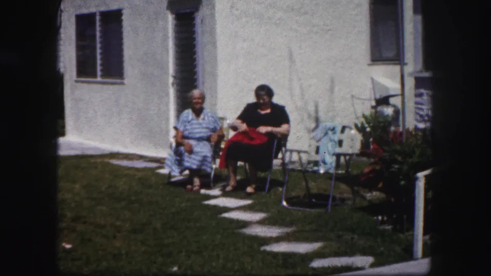 Two older women sit in lawn chairs in a Tampa backyard, one holding a red cloth, with patio furniture and house windows nearby.