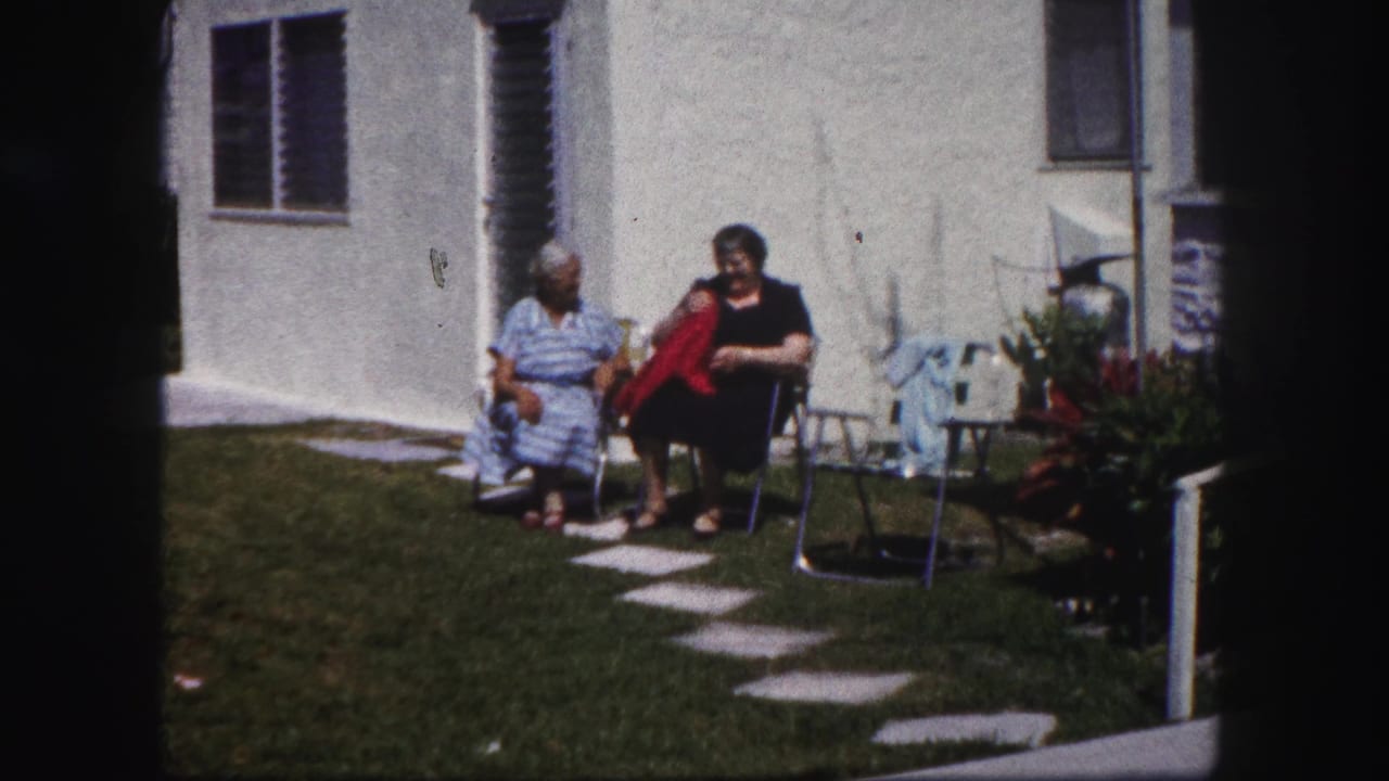 Two older women sit on folding chairs in a sunny yard beside a white stucco house.