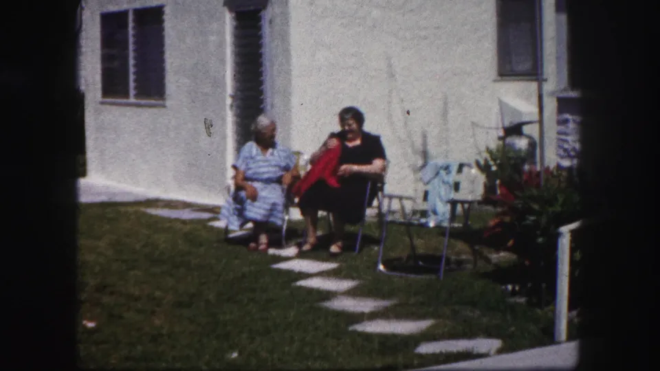 In 1960 Tampa, two older women sit outside on folding chairs near lawn pavers and a white stucco wall.