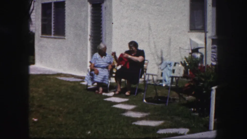 Two older women sit in a Tampa backyard, one handling a red garment while the other faces her in conversation.