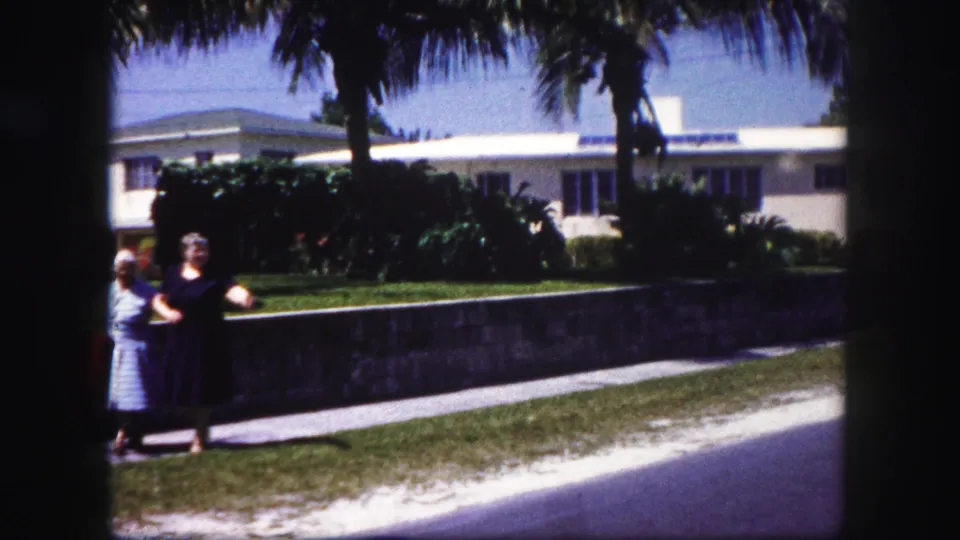 Two adult women walk arm in arm in a 1960 Tampa residential street scene, with a low wall, palms, lawn, and white house behind them.