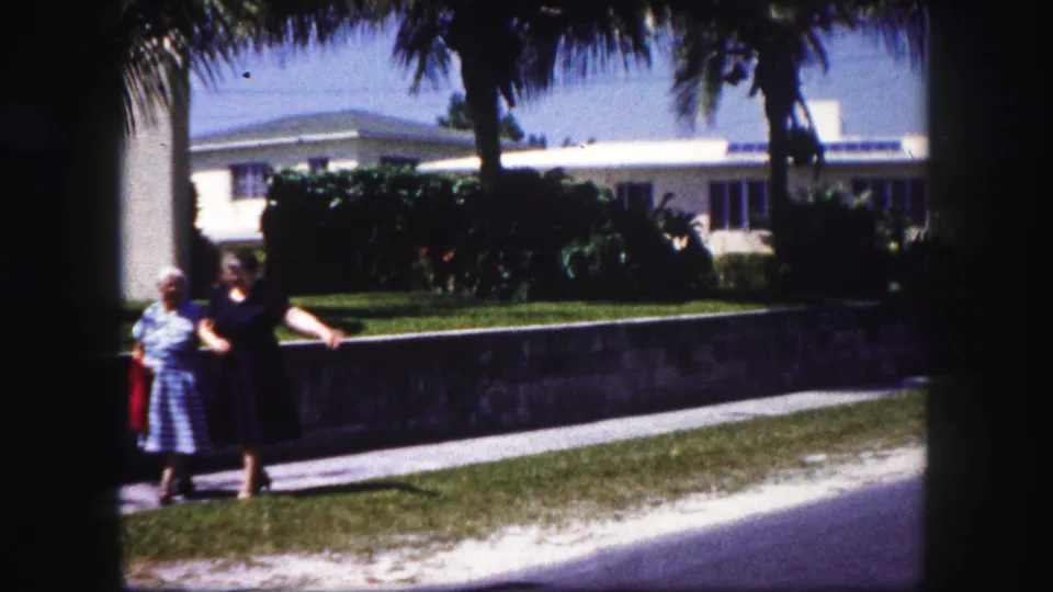 Two women in period dresses stand near a Tampa sidewalk in 1960 home-movie footage, framed by palms, shrubs, a low wall, and a white house.