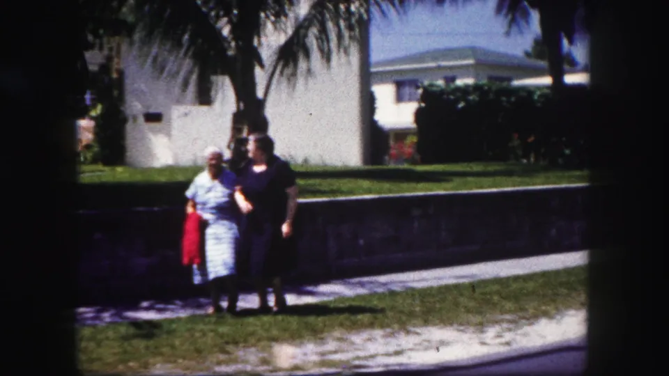 The two women continue along the Tampa sidewalk, one in a light striped dress with a red jacket and the other in a dark dress.