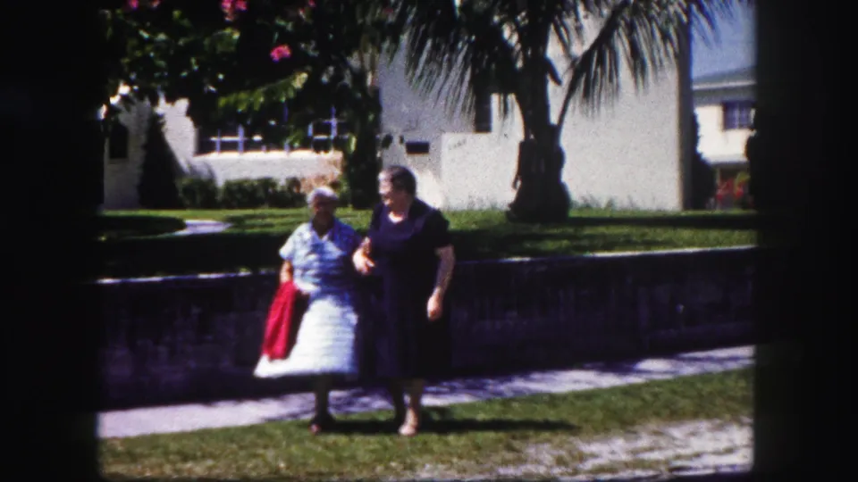 The final frame shows the two older women walking arm in arm, with the red jacket visible against the lawn, wall, palms, and Tampa residential backdrop.