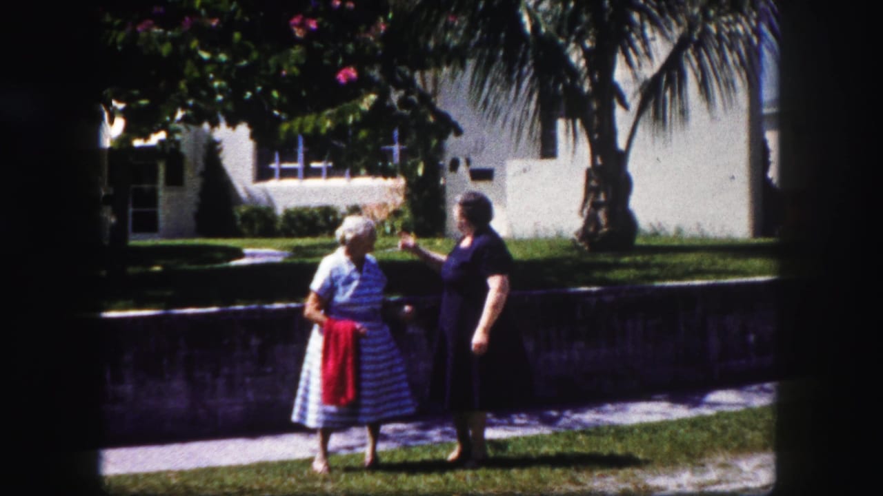 Two women stand on grass near a sidewalk and low wall, with palms and a pale house behind them.