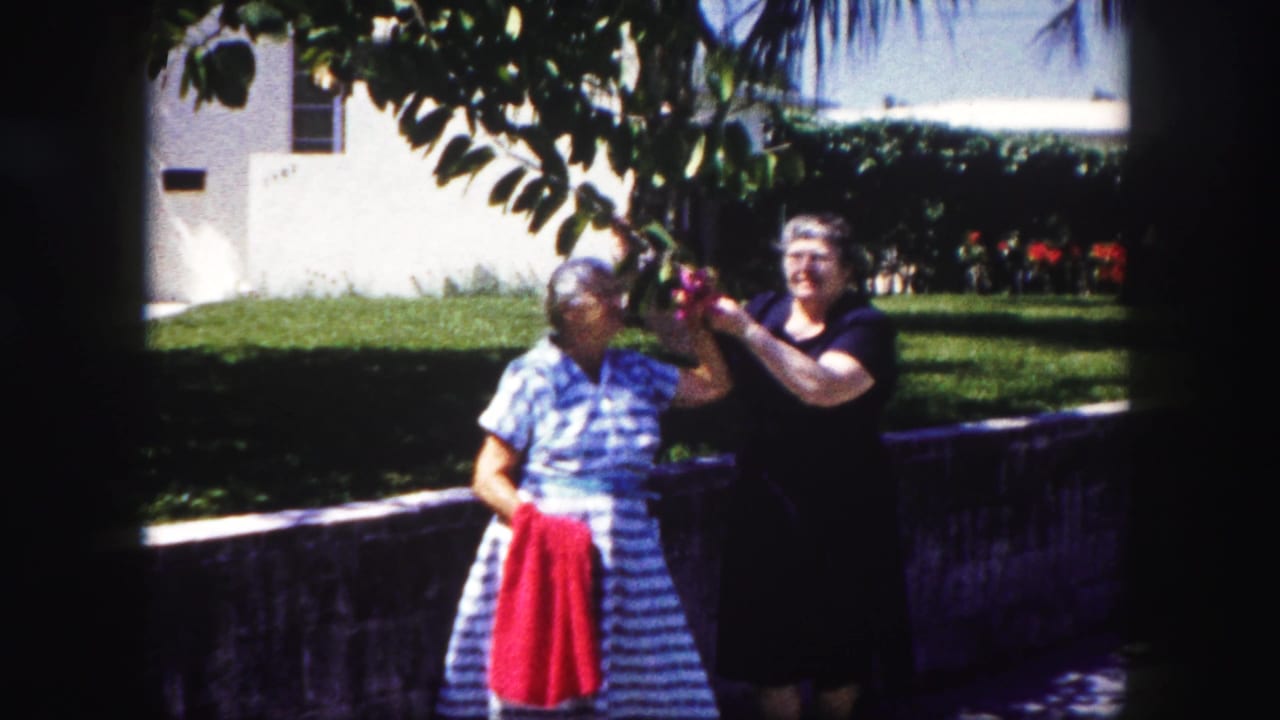 In 1960 Tampa home-movie footage, two adult women pose in a sunny yard as one holds a red towel and the other reaches toward foliage.