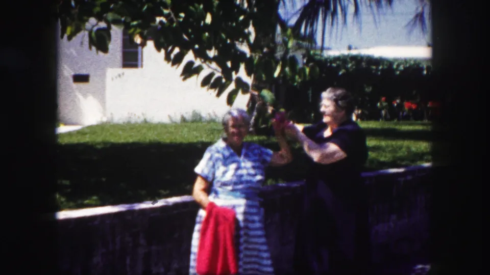 Two adult women in a Tampa yard appear to laugh and pose as one woman holds a red towel and the other touches a tree branch.