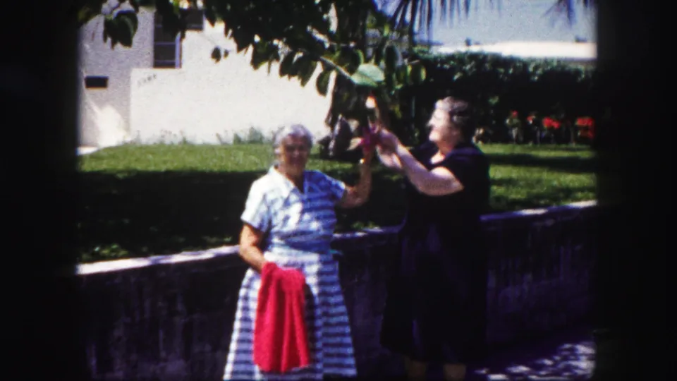 At the end of the clip, the woman in dark clothing looks up toward foliage while the other stands in front with a red towel.