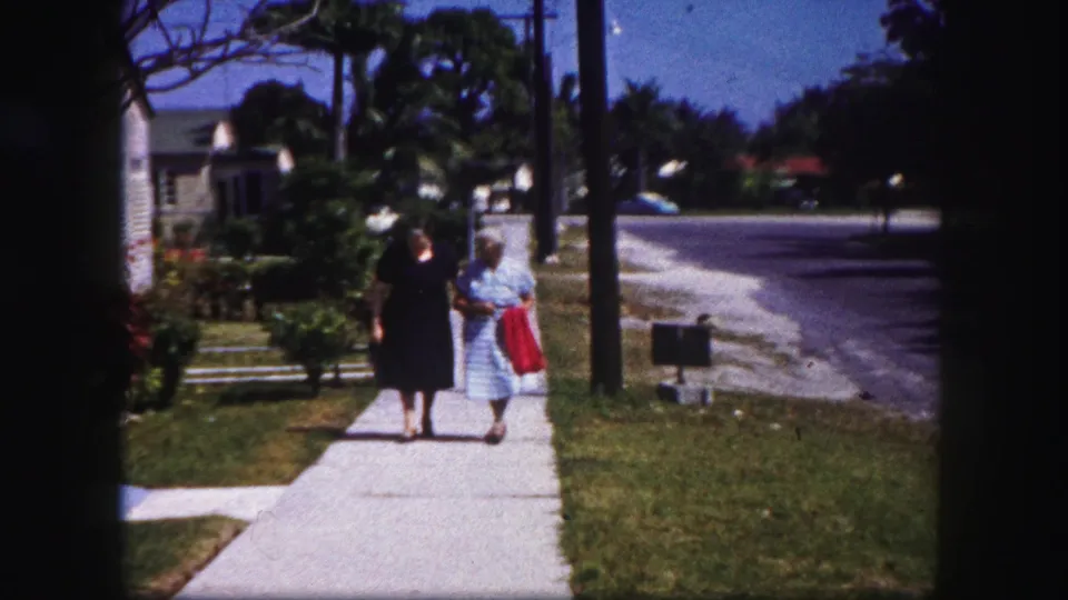 The pair continues toward the camera on a sunny 1960 Tampa sidewalk, one in a striped dress carrying a red cloth.