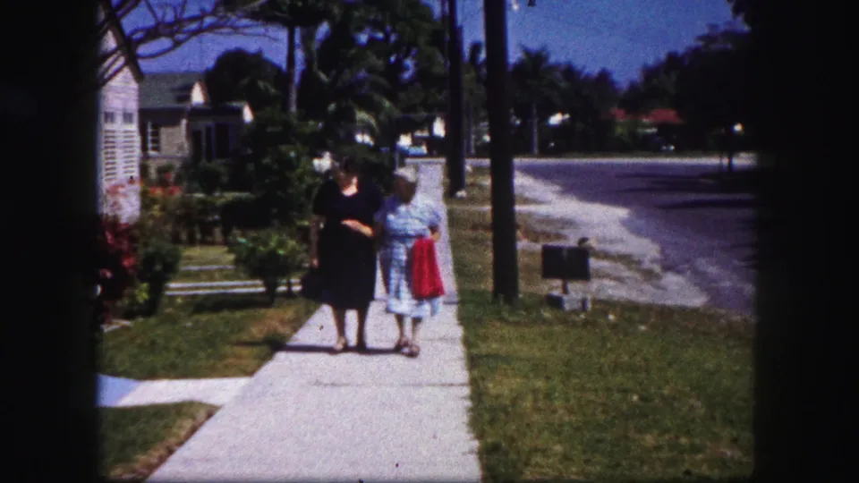 Near the end of the shot, the two older women appear to slow on the Tampa sidewalk, framed by lawns, houses, palms, and street edge.
