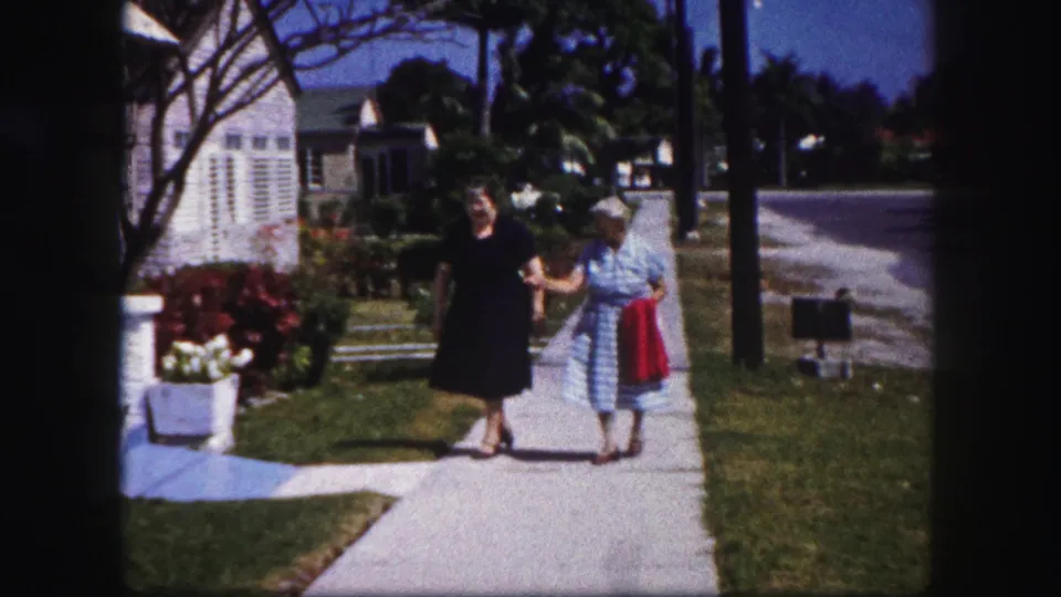The women walk together in a sunny Tampa residential setting, with one wearing a black dress and the other holding a red cloth.
