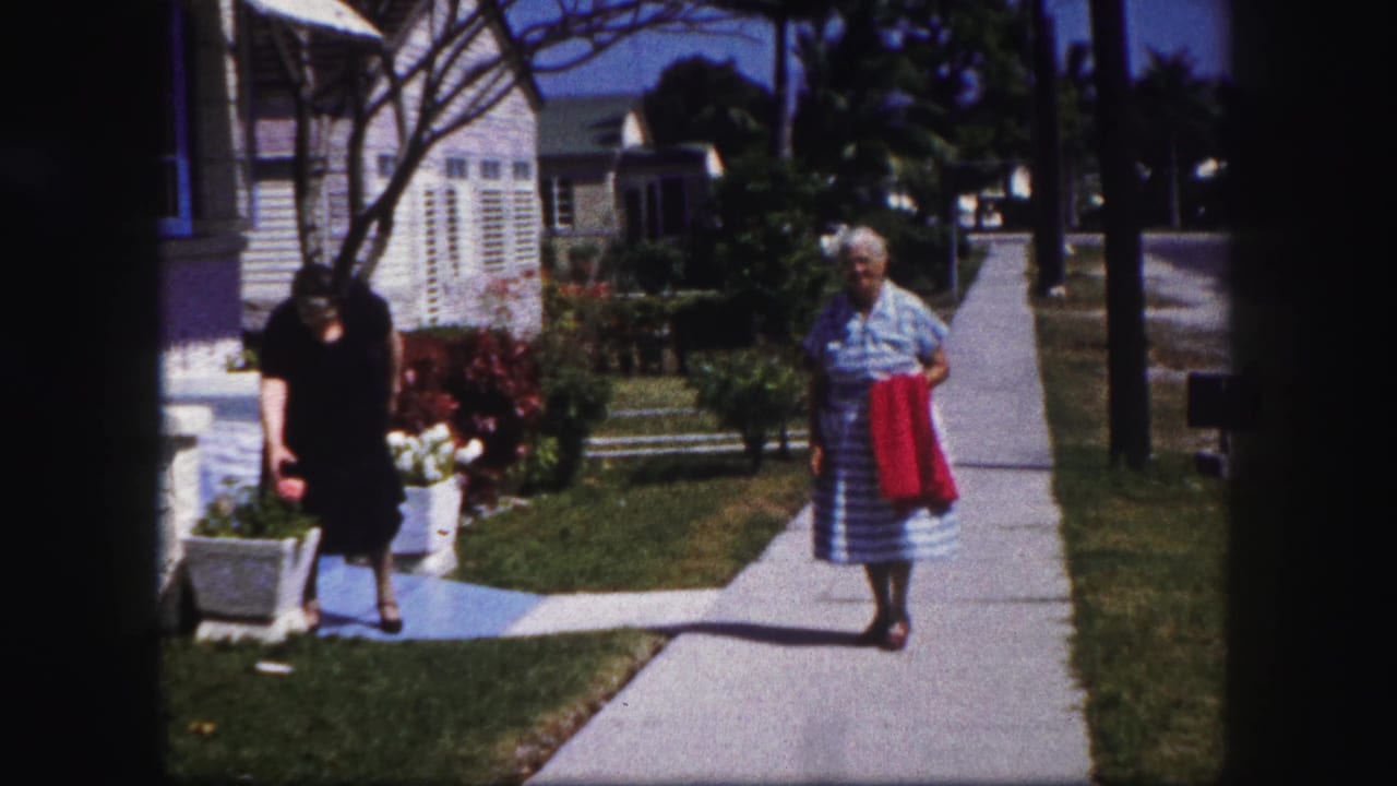 Two women stand near a sidewalk and porch outside a suburban house.