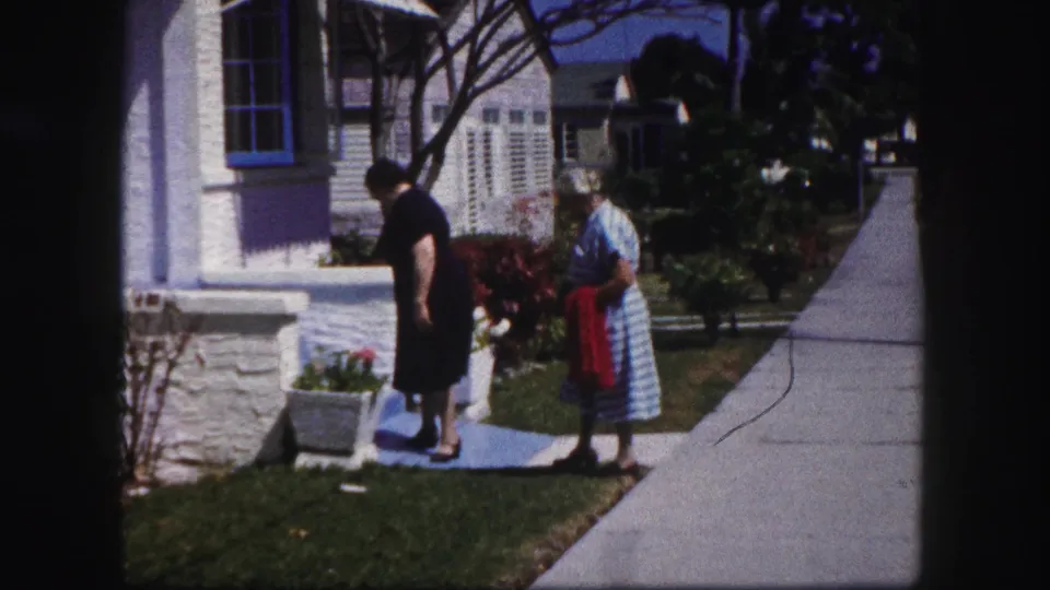 The two women pause near the white porch steps in a sunny Tampa suburban yard, with planters and a sidewalk visible.