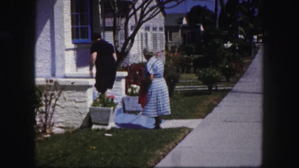 A woman in black bends toward a white porch planter as the older woman stands nearby in the residential Tampa yard.