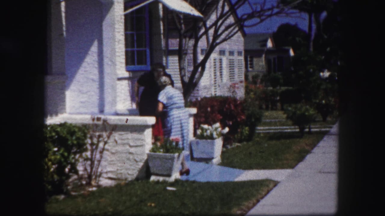 A 1960 Tampa home-movie frame shows one woman in a striped dress near another at the porch edge of a white house.