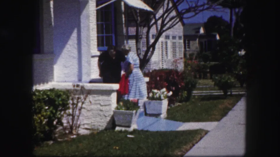 Two women interact beside the porch railing of a Tampa suburban house, with lawn, shrubs, and potted flowers visible.