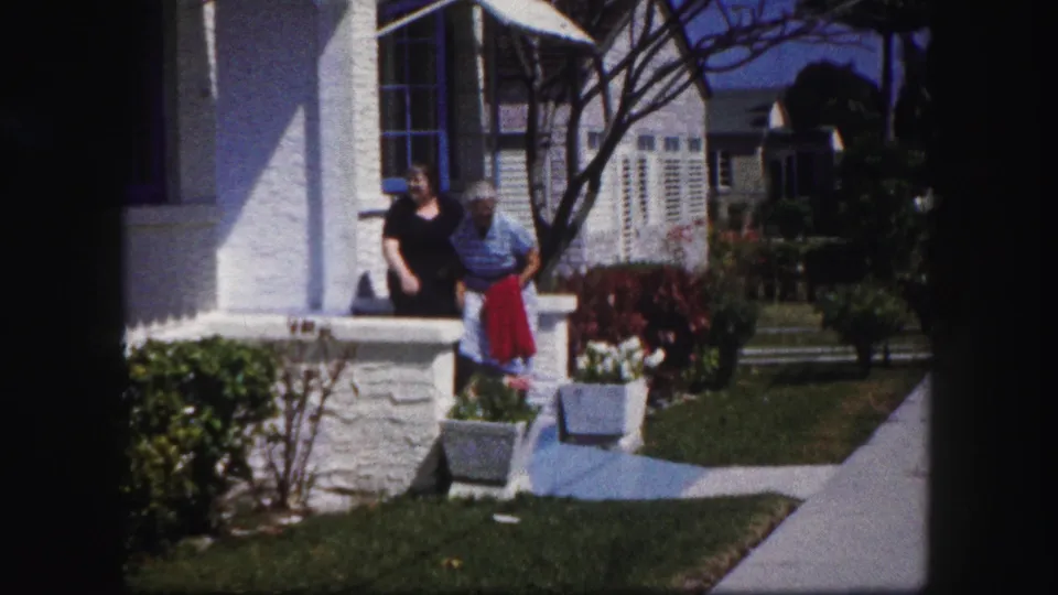 At the end of the 1960 Tampa porch scene, a woman holds a red garment while another woman stands near the white stucco wall.