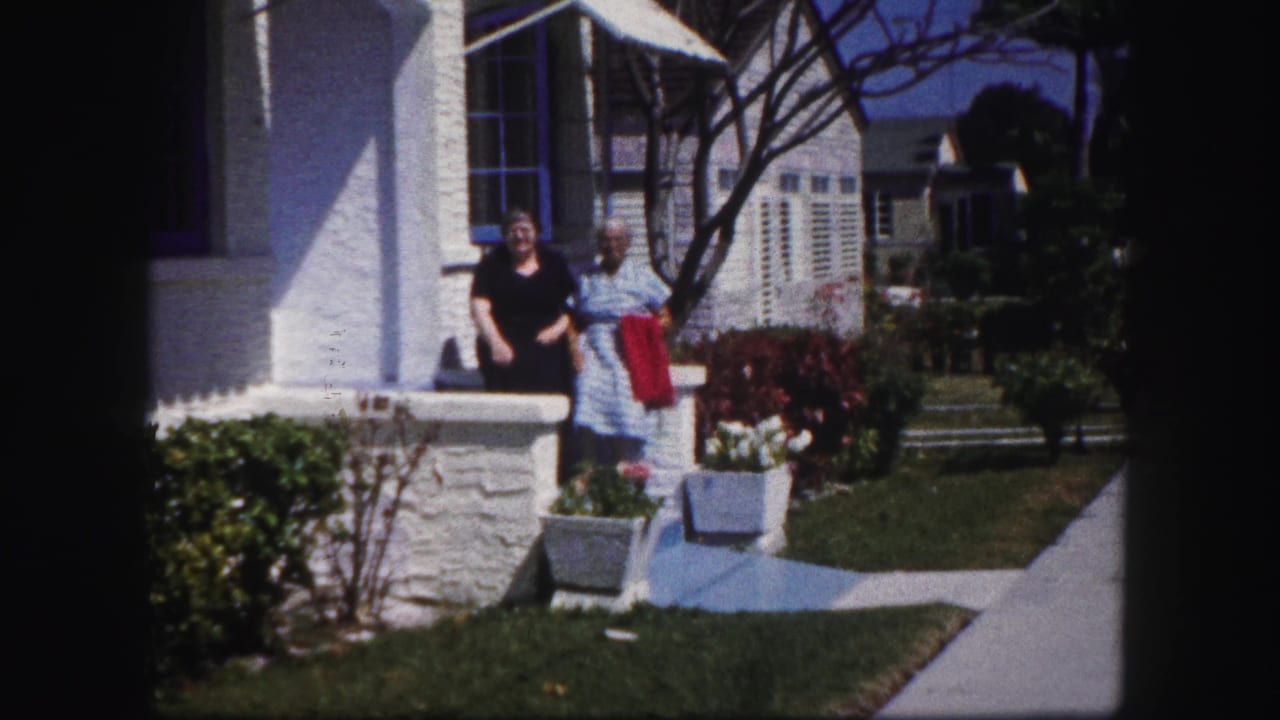 In 1960 Tampa home-movie footage, two older women appear near a white stucco house with blue-trimmed windows and a sunny sidewalk.
