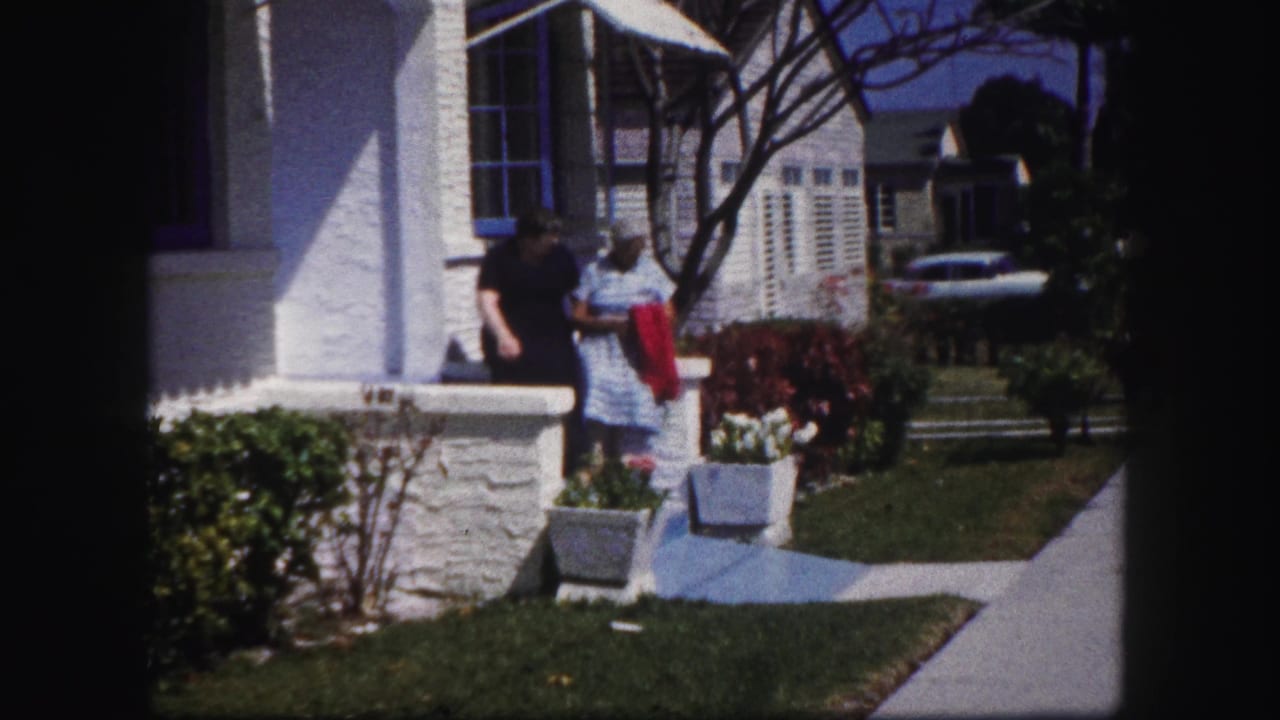 Two older women move along a residential sidewalk in Tampa, with a white house, shrubs, and parked car partly visible in the background.
