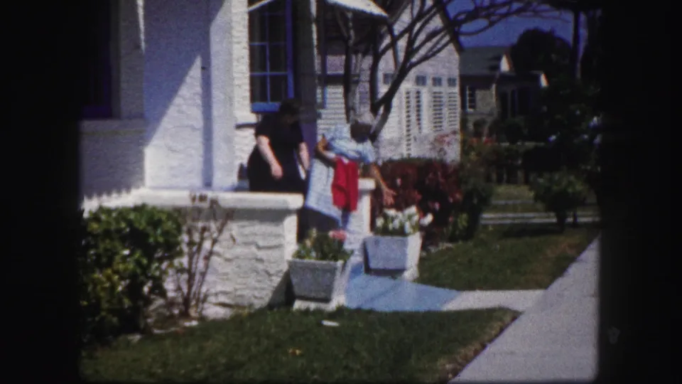 A 1960 Tampa home-movie frame shows two older women beside a white stucco porch, with flowers, lawn, and a bright sidewalk in view.