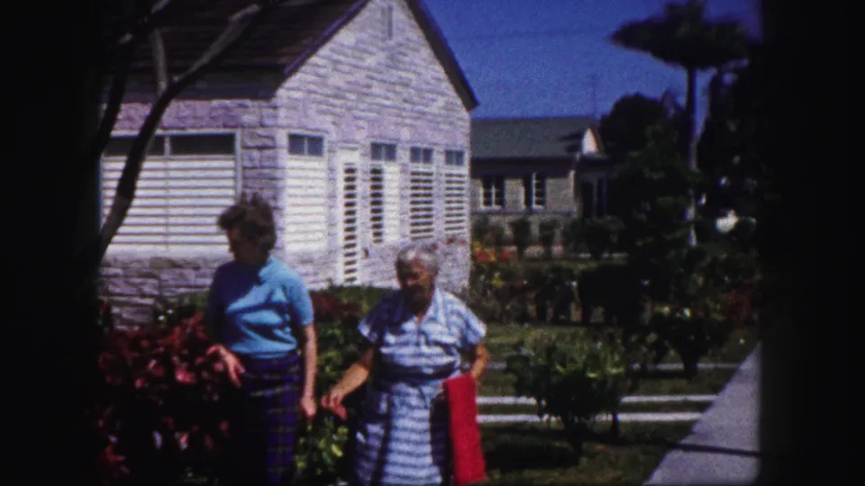 Two older women walk side by side in a Tampa residential yard, with a white house, garden plantings, and palm-like trees visible behind them.