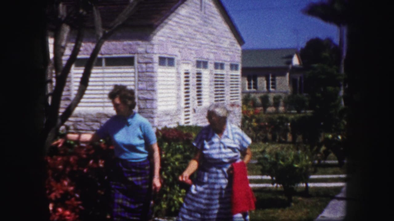 In Tampa, Florida circa 1960, two women pass a red flower bed near a pale house with white louvered shutters.