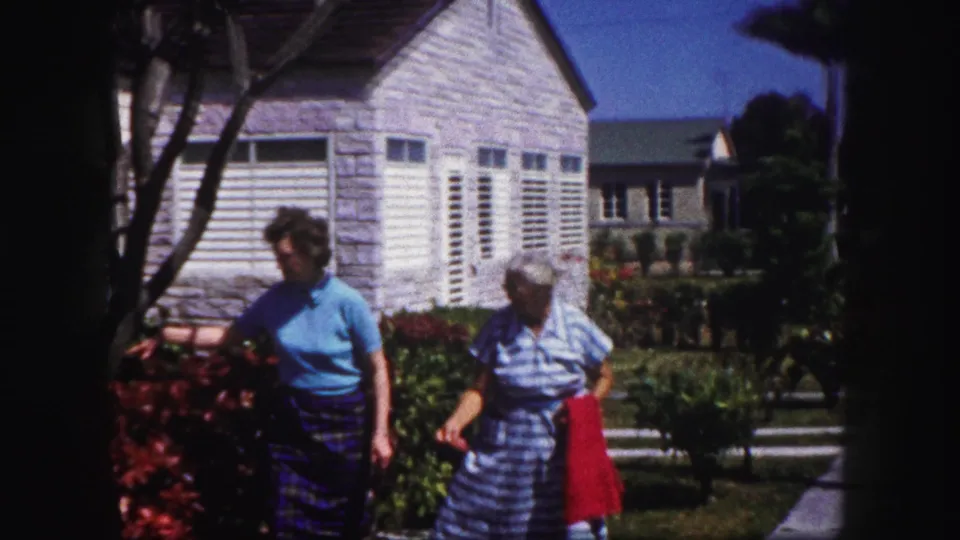 In Tampa, Florida circa 1960, two women pass a red flower bed near a pale house with white louvered shutters.