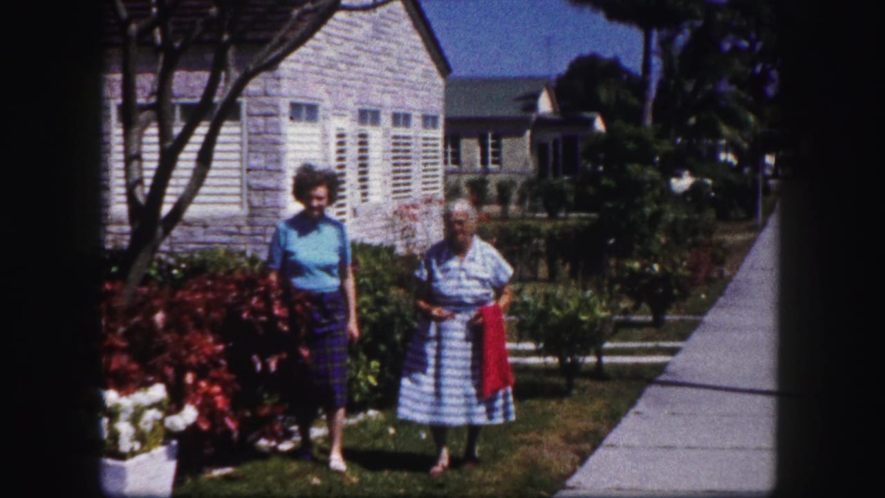 Two women in a Tampa garden pause or slow near shrubs, with the shuttered house and concrete sidewalk visible behind them.