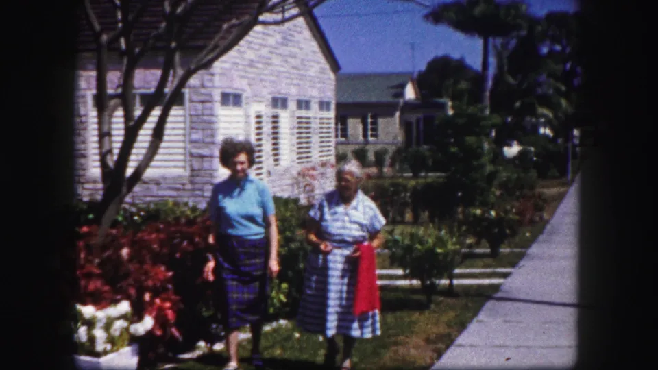 The younger and older women continue along the garden edge, passing red and white flowers in the circa-1960 Tampa home-movie scene.