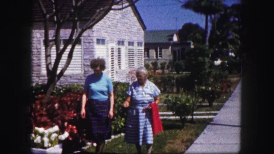 Two women walk side by side through a sunny Tampa garden, framed by flowers, shrubs, sidewalk, and a pale shuttered house.